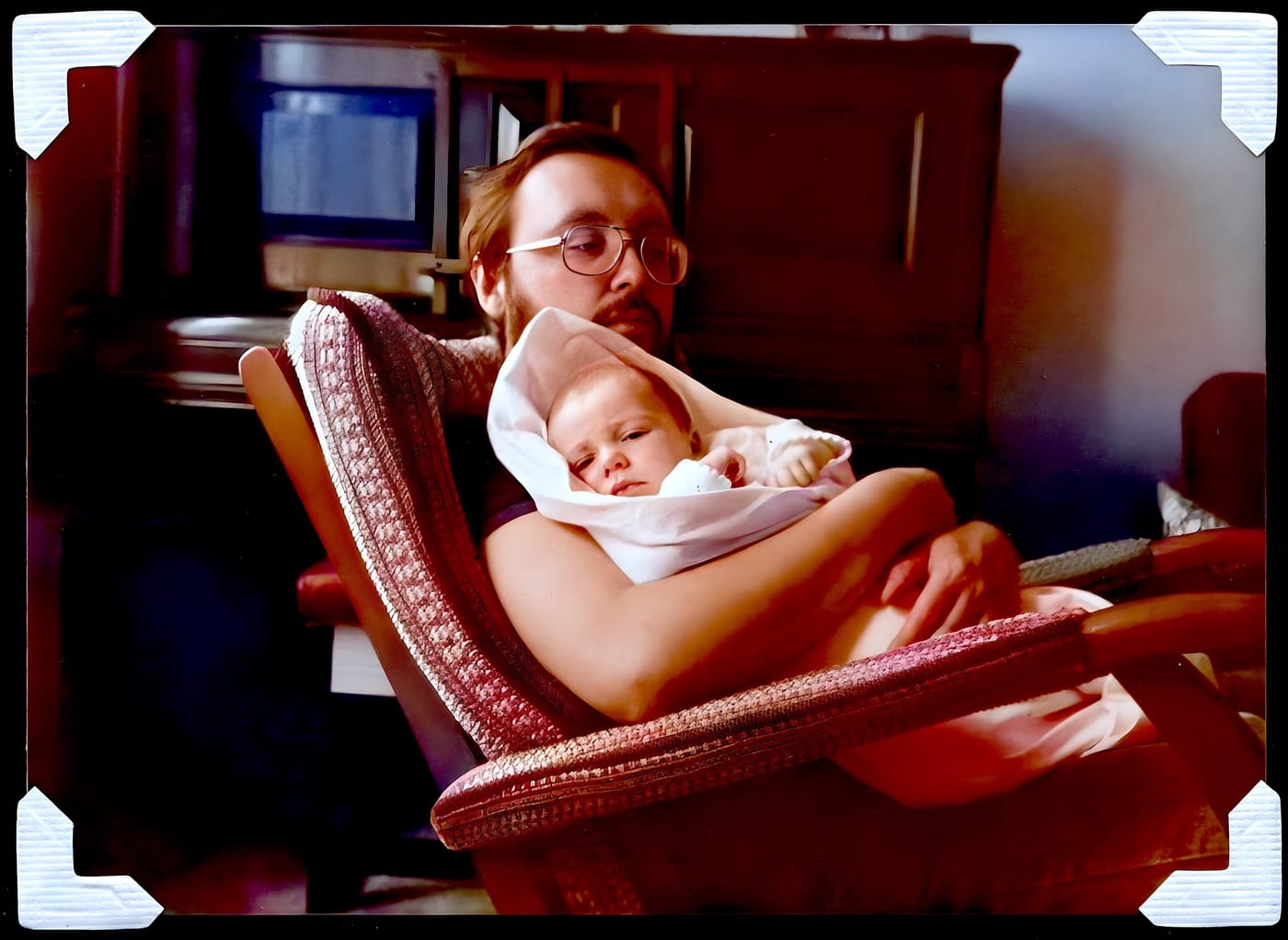 A man in glasses holding a sleeping baby, photographed indoors in the 1970s